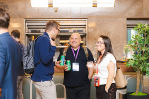 Three colleagues chat at a bar area; two men hold drinks while a woman in glasses smiles, all wearing conference badges and lanyards.