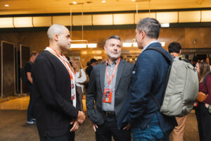 Three men in business attire chat at a conference, each wearing name badges and lanyards in a bustling networking area