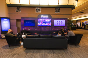 Roku demo lounge with black sofas and people seated in front of a purple-lit display wall and screens labeled Roku.