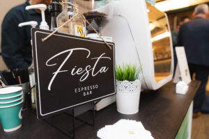 Black sign reads'Fiesta Espresso Bar' hanging on a cafe counter with cups, pump bottles, and a small white plant pot in the background.