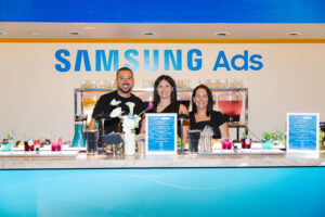 Three smiling staff members stand behind a cocktail bar at a Samsung Ads event, with colorful drinks on the counter and a blue Samsung Ads sign behind them.