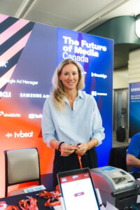 Woman in a light-blue shirt stands at a conference booth with a'The Future of Media Canada' banner behind her.