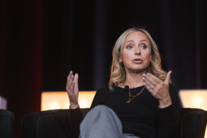 Blonde woman in a black top sits on a stage, speaking with a lapel mic and using expressive hand gestures.