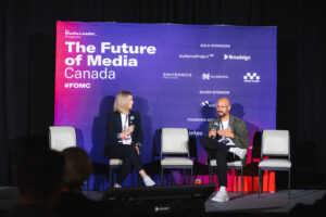 Two panelists sit on stage in front of a purple banner reading'The Future of Media Canada' during a conference, each holding a microphone and talking to an audience.