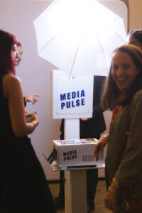 People at a media booth with a bright umbrella light and a sign reading'MEDIA PULSE' on a white pedestal.