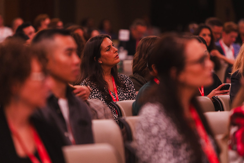 Audience of conference attendees seated in rows, a woman in a patterned blouse in the center listening attentively while others around her look on.