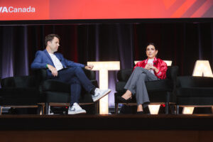 Two panelists seated on stage in black chairs, a man in a blue blazer and white sneakers speaking while a woman in a red jacket listens; large illuminated letters backdrop.