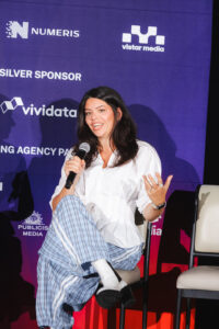 Woman in a white blouse and blue striped pants sits on stage, speaking into a microphone with her left hand raised.
