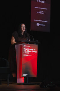 Woman presenting at a podium on stage about TV advertising; banner reads'The Future of TV Advertising Canada #FTVACanada' with sponsor board in the background
