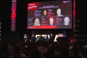 Panel discussion on a stage at a conference, six speakers seated in armchairs facing an audience, large screen behind lists panelists' names and topics.