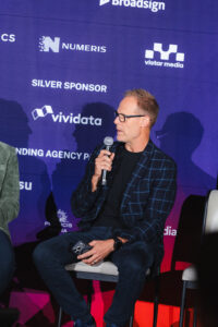 Man in a checkered blazer speaks into a microphone on stage at a conference backdrop with sponsor logos behind him.
