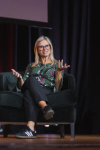 Woman with blonde hair and glasses seated on a stage in a dark chair, gesturing with her hands as she speaks in a panel setting.