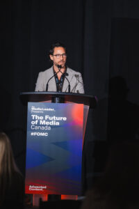 Male presenter at a lectern with a microphone, banner reads The Future of Media Canada (#FOMC). Readable stage lighting highlights the speaker in a gray blazer and glasses.