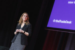 Female presenter on stage in a black blazer and patterned dress, with a purple screen displaying theTradeDesk logo.