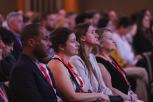 Dair of diverse conference attendees seated in an auditorium, wearing red lanyards, watching a presentation attentively.
