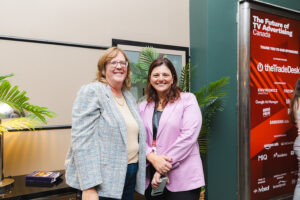 Two women posing together at a conference, smiling, with plants and a red event banner in the background.