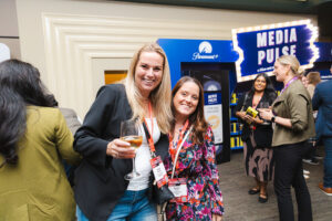 Two smiling women posing for a photo at a media event, one holding a glass of wine, with a Media Pulse backdrop in the background.