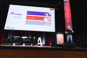 A man speaks on a stage beside a large projection screen showing a Vividata slide with three colored bars labeled Truth, Vividata Spatial, and Vividata Relevance, and a few chairs on stage in the background.
