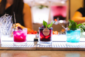 Three colorful cocktails on a bar counter, garnished with herbs and berries; middle glass has a Samsung Ad label.