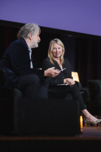 Two panelists on stage in conversation: a gray-haired man with a beard and a smiling blonde woman, holding glasses.