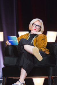 Woman with blonde bob and black glasses sits on a black armchair on stage, resting her chin on her hand and holding a blue pamphlet.