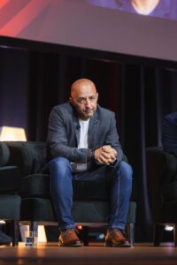 Man with a shaved head sits on a dark-green armchair on a stage, hands clasped, wearing a gray blazer, white shirt, jeans, and brown shoes.
