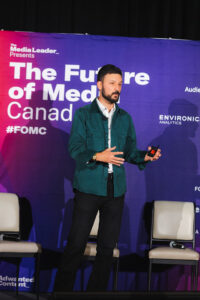 Male presenter in teal jacket speaks on stage at a media conference, backdrop reads “The Future of Media Canada.”