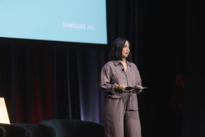 Female presenter on a stage, holding notes and microphone, with a Samsung Ads screen behind her.