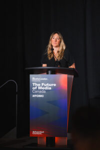 Woman speaking at a podium on stage for The Future of Media Canada event; banner shows #FOMC and ‘The Future of Media Canada.’