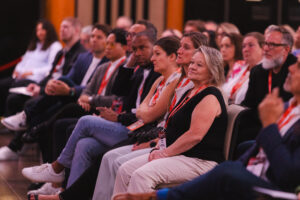Audience seated in a conference hall attentively listening to a speaker, with lanyards and badges visible in the foreground.