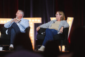 Two panelists sit in armchairs on a stage, talking during a panel discussion with warm backlighting and large illuminated letters in the background.
