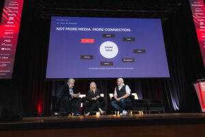 Three panelists seated on a lit stage in front of a large screen that reads'NOT MORE MEDIA. MORE CONNECTION.' with a central circle labeled 'SHARK NINJA' and surrounding labels like TV, DIGITAL, SEARCH, RETAIL.