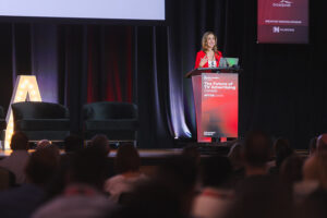 Woman in a red blazer speaks at a conference podium on a dimly lit stage with a banner that reads'The Future of TV Advertising Canada'.