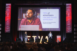 Conference stage with a man wearing headphones presenting beside a large screen showing the slide:'78% of advertisers use last-touch attribution'. Audience seated in front of the stage.