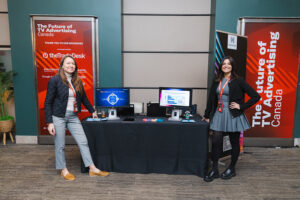 Two women stand at a conference booth with two computer screens and banners about TV advertising in Canada.
