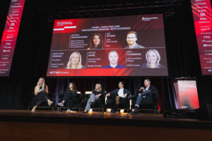 Panel discussion at a conference with five speakers seated on stage; a large screen behind them shows headshots and bios of the panelists and the event branding.