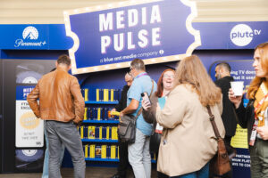 People gather at a Media Pulse booth with bright blue signage (Paramount+ and Pluto TV logos) and shelves of yellow items in the background, at a convention.
