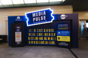 Illuminated MEDIA PULSE sign atop a blue trade show booth with shelves of yellow boxes nearby.