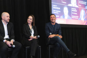 Three panelists seated on stage at a conference, each holding a microphone and smiling, with a projection screen behind them.