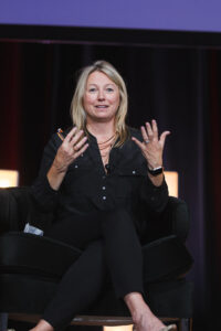 Woman speaker seated on a stage, gesturing with hands while talking into a mic, dark backdrop behind.