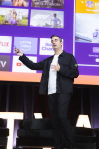 Male presenter on stage gesturing with a remote, backdrop shows a purple screen with streaming service logos (Netflix, Prime Video) and audience seating area behind him.
