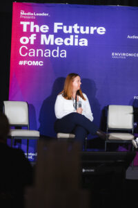 Woman seated on stage holding a microphone during a media conference with a purple backdrop reading'The Future of Media Canada' and the hashtag #FOMC
