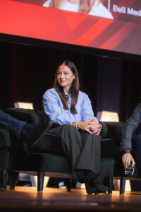Panelist woman in a light blue shirt sits on stage with folded hands during a conference panel.
