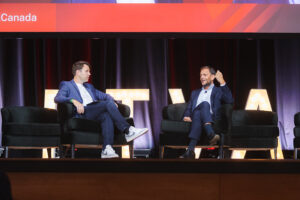 Two men in suits sit on stage in armchairs during a panel discussion, with large lit letters behind them and a red banner showing'Canada' above.