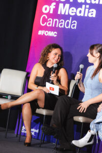 Two women on a stage panel sit in chairs; one speaks into a microphone and holds a card with a YouTube play icon while the other chats, against a purple backdrop that reads'Canada' and '#FOMC'.