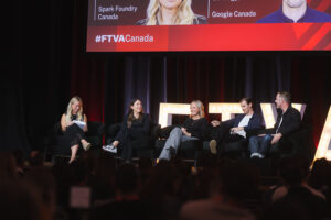 Five panelists sit on stage in black chairs during a discussion, with a large, lit backdrop and an audience visible in the foreground.