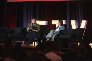 Two women on stage in armchairs having a discussion, audience visible in the foreground, large illuminated letters behind them.