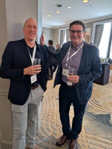 Two men in business casual attire at a conference reception, smiling and holding drinks with conference badges visible.