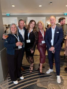Group of five adults at a social event posing for a photo in a hotel corridor, each with a name badge and drinks in hand.