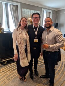 Three smiling adults posing for a photo at a formal event, wearing lanyards, in an elegant room with a bar and chandelier in the background.